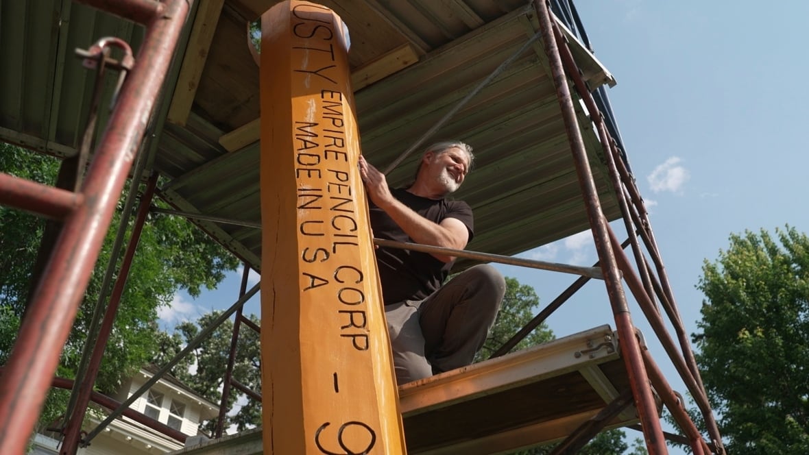 A bearded man perches on scaffolding and touches a giant pencil with the words "EMPIRE PENCIL CORP MADE IN USA" carved into the side.