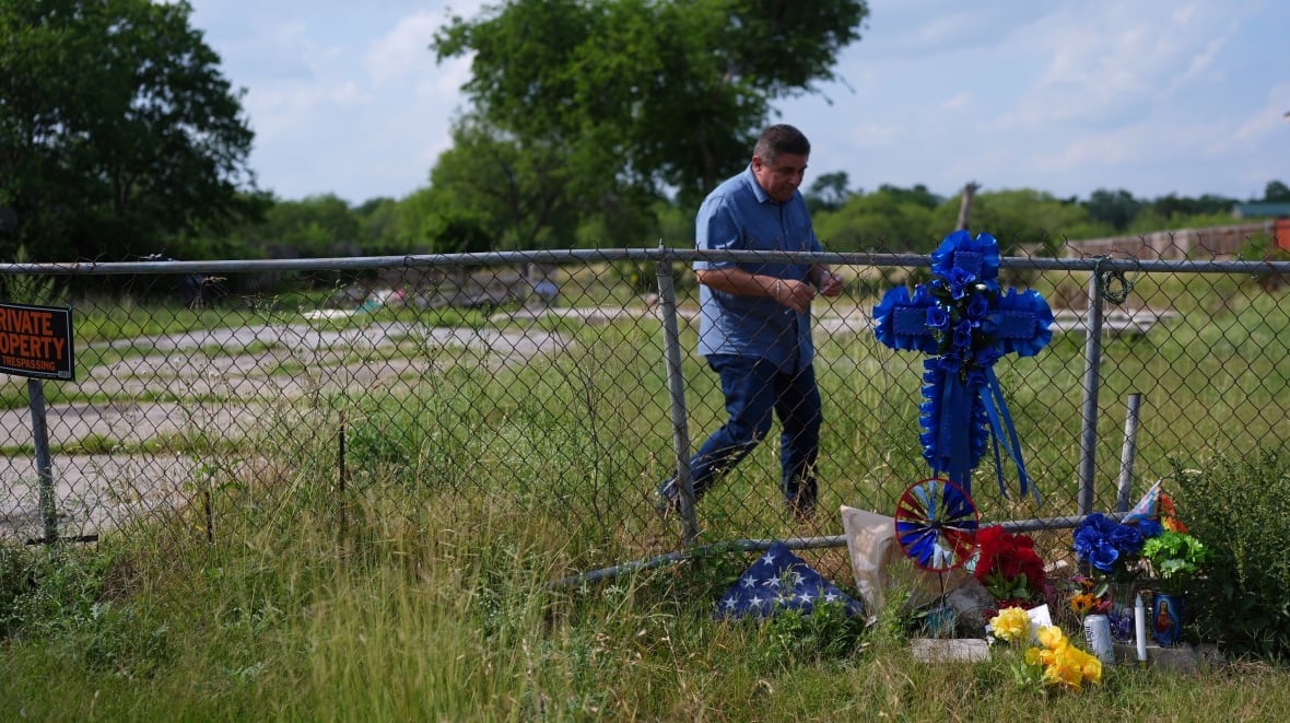 A man approaches a memorial near a wire fence, with flowers at the bottom of the fence and a large blue cross attached to the fence itself. The fence crosses a green, overgrown lawn or field, and a structure of some type is visible in the background.