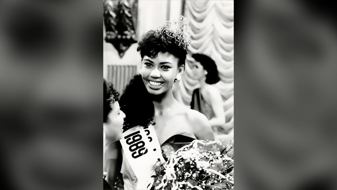 A Black woman, with long curly black hair, smiles as she poses with a crown on her head, a white sash with "1989" on it hanging over her shoulder, and a bouquet of flowers in her arms.