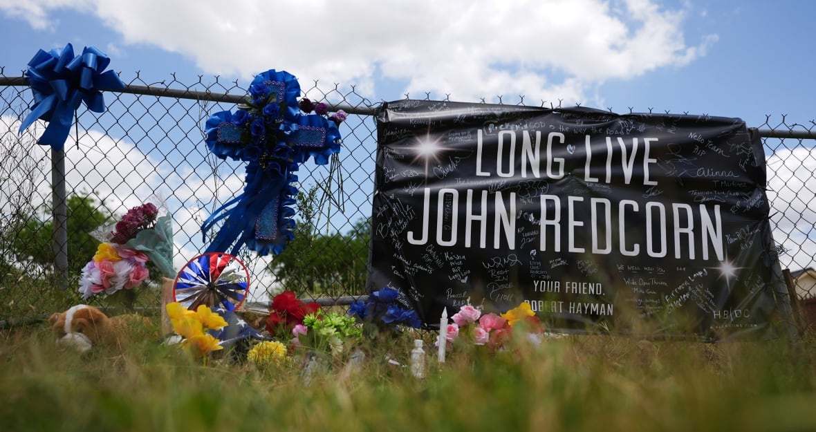 A sign hangs from a fence that says "Long Live John Redcorn" in large letters, with "Your Friend Robert Hayman" underneath. It has been hand-signed many times. Beside it, a large blue cross hangs from the fence, and a number of flowers and flags rest at the bottom of the fence in the grass.