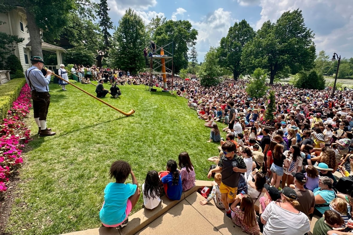 A crowd of people on a sunny day sit and stand on the street in front of a lawn featuring a giant pencil surrounded by scaffolding. People dressed in old-timey clothes stand on the lawn and blow into long horns.